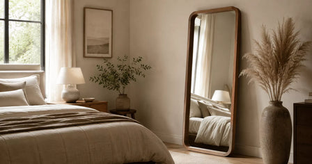 Tall full-length wooden framed mirror placed against a warm neutral bedroom wall beside a bed, styled with natural daylight, soft beige furnishings and minimal decor.
