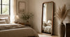 Tall full-length wooden framed mirror placed against a warm neutral bedroom wall beside a bed, styled with natural daylight, soft beige furnishings and minimal decor.
