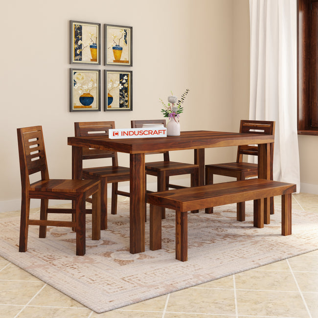 A dining bench set with a solid wood Sheesham bench and upholstered fabric chairs, arranged around a rectangular dining table, placed on a rug in a room with framed artwork on the walls and a vase with flowers on the table.