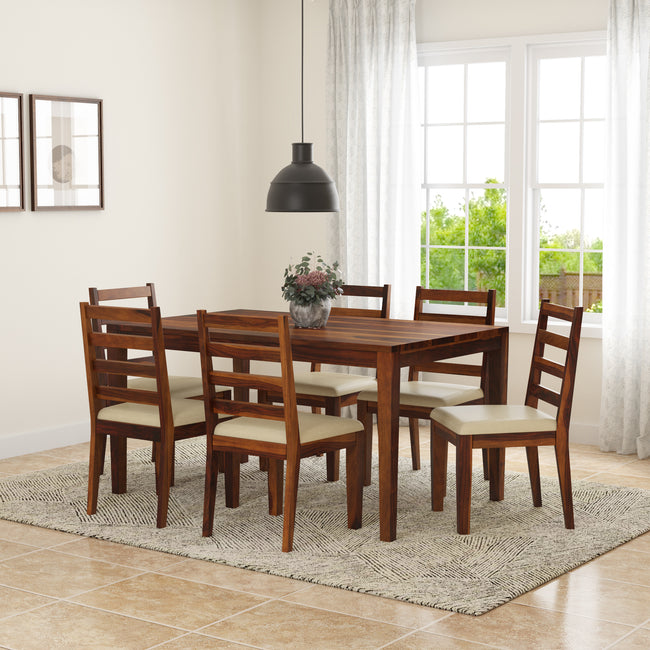 A wooden dining set comprising a rectangular table and six chairs with beige cushions, placed in a room with a window, a pendant light, and a floral arrangement on the table.
